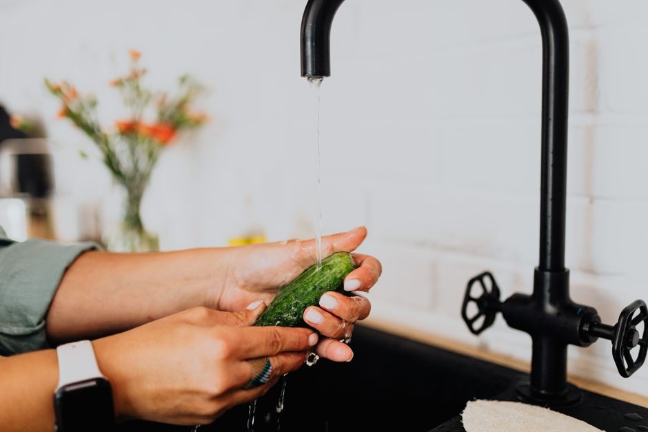 Hands washing a fresh cucumber under running water in a kitchen setting.
