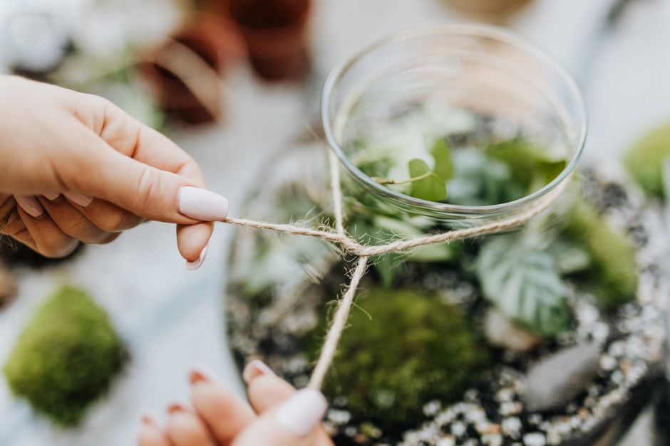 Detailed view of hands tying jute rope around a glass terrarium with green plants.