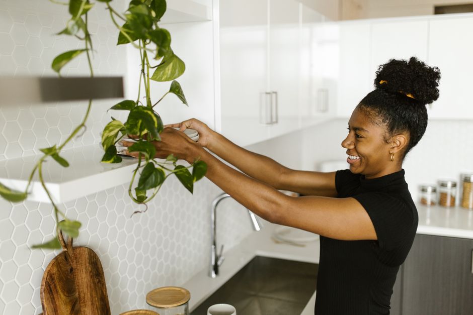 A woman joyfully arranges a hanging plant in a bright modern kitchen with white hexagonal tiles.