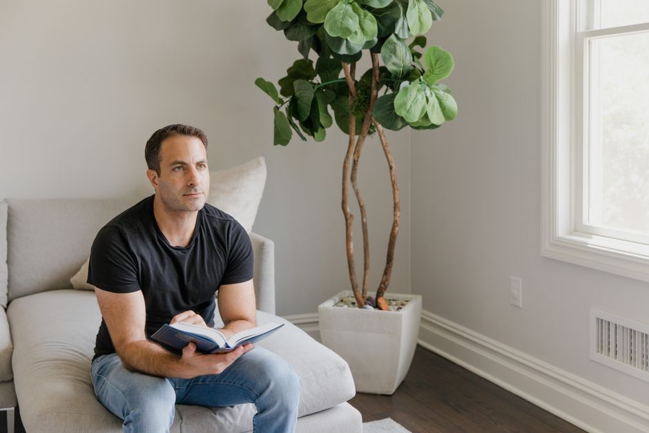 A thoughtful man reading a book on a couch near a window at home.