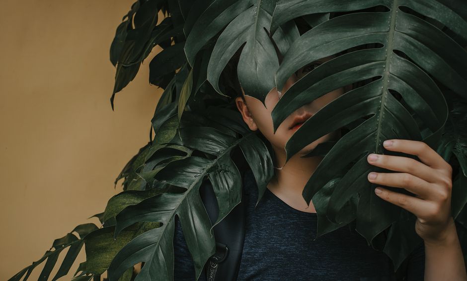 A young man partially hidden behind large tropical leaves, evoking tranquility and mystery.