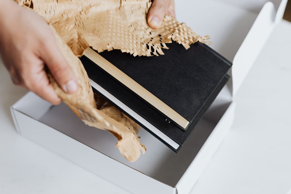 From above crop adult male placing black notebooks with craft perforated paper in white cardboard box while preparing ma